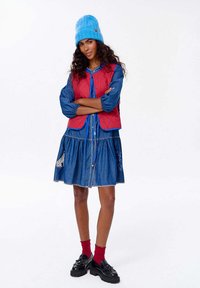 Červená prešívaná vesta cez modré denimové šaty s viacúrovňovou sukňou, ktorá má kontrastné stehy a dekoratívne prvky. Modrá pletená čiapka a čierne topánky.