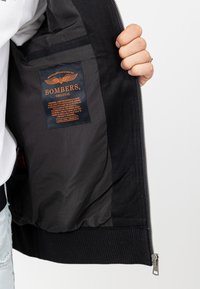 Zwarte bomberjack met een ritsluiting, ribbelmanchetten en een binnenlabel met "BOMBERS ORIGINAL" in oranje en blauwe tekst.
