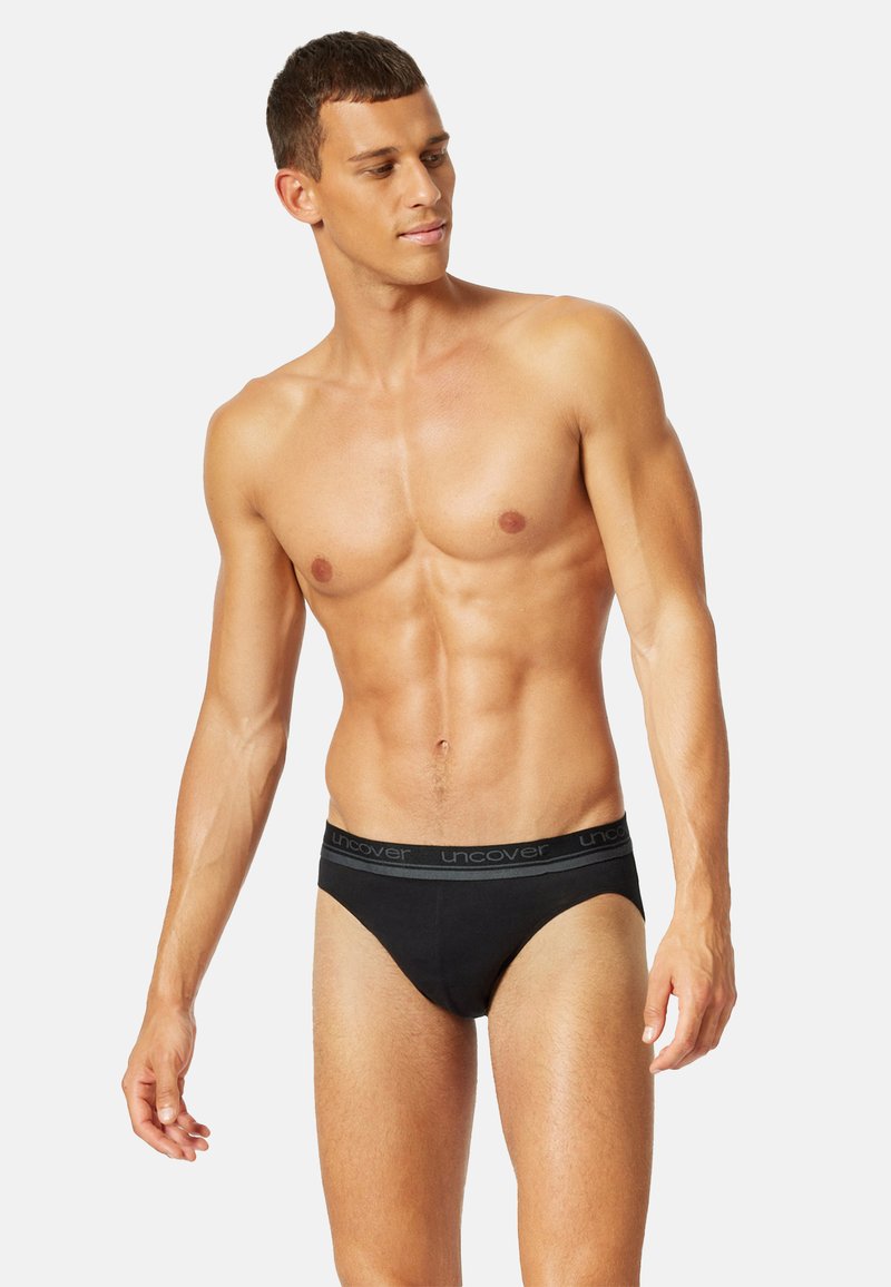 Muskulöser Mann mit kurzen Haaren, der schwarze Slips mit einem "uncover"-Bund trägt, steht und nach links schaut, vor einem schlichten Hintergrund.