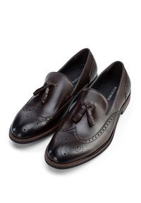 Los mocasines de cuero marrón presentan un diseño brogue con perforaciones y borlas en la parte delantera. Los zapatos tienen una forma elegante y una superficie texturizada.
