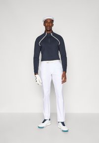 Pulôver azul marinho de manga longa com fecho éclair e detalhes brancos, combinado com calças brancas e sapatos de golfe. Os acessórios incluem uma capa branca e uma luva.