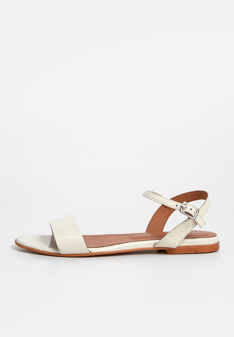 Sandalia plana con una amplia correa de cuero blanco, correa en el tobillo ajustable, plantilla de cuero marrón y suela delgada de goma color tostado. Diseño minimalista.