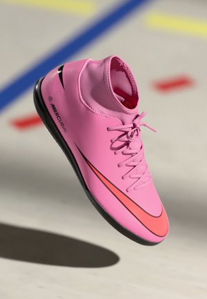 Chaussure de football d'intérieur Nike Mercurial rose avec semelle noire et swoosh orange vif, présentée devant un fond flou de terrain de sport.