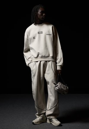 Person mit dunkler Hautfarbe und Dreadlocks trägt ein beiges Athleisure-Set mit schwarzer Paspelierung und hält eine kleine Camouflage-Tasche vor schwarzem Hintergrund.