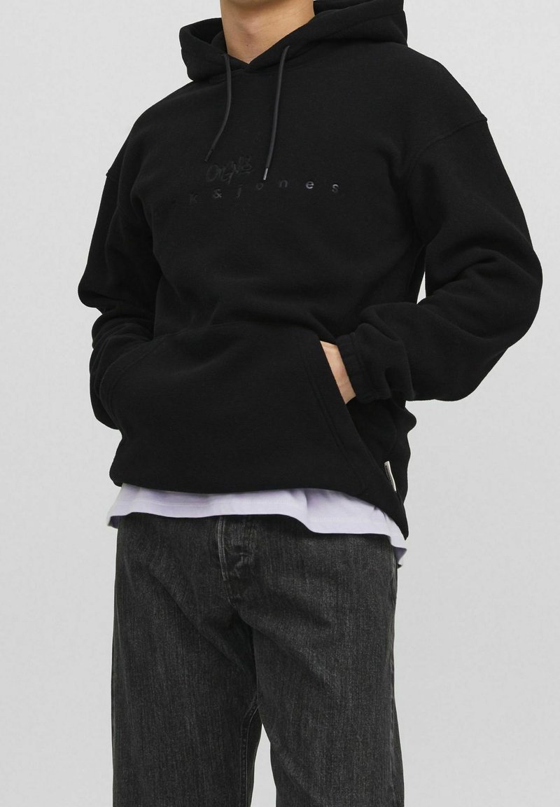 Zwarte hoodie van zachte stof, met een voorzak, een verstelbare trekkoordkap en een subtiele logo op de borst. Draagt over een wit shirt.