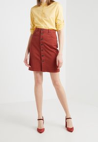 Röd denimkjol med dekorativa knappar framtill och två fickor, matchad med en ljusgul skjorta med krage och röda högklackade skor.