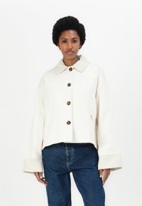 Cream-colored cropped jacket with a pointed collar, brown buttons, and wide cuffs. Worn over blue denim jeans. Smooth texture.