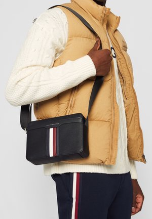 Mann trägt eine beige Steppweste und einen cremefarbenen Zopfpullover, trägt eine schwarze Umhängetasche mit roten und weißen Streifen.