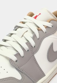 Tênis brancos com detalhes cinzas, apresentando cadarços brancos, um swoosh branco em destaque e um pequeno logotipo vermelho na língua.