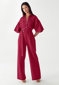 Moteris stovi su rankomis už nugaros, vilkėdama raudoną diržinį kombinezoną su pūstomis rankovėmis ir smėlio spalvos atvirais sandalais prieš paprastą baltą foną.