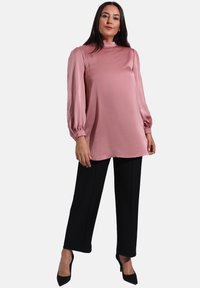 Blusa a maniche lunghe in morbido tessuto rosa chiaro con collo alto e maniche gather, abbinata a pantaloni neri su misura e tacchi a punta.