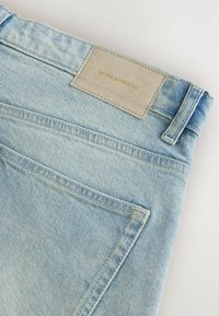 Jeans din denim de culoare bleu deschis, cu o suprafață texturată, având un eticheta din piele bej care spune "DENIM SUPPLY Co." și un design standard pentru buzunarul din spate.