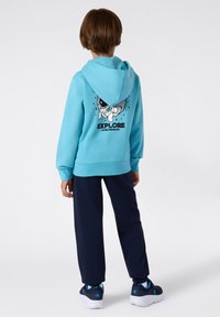 Hoodie turchese con cappuccio, caratterizzata da una grafica di un esploratore spaziale e la scritta "ESPLORA OLTRE LA LUNA" sul retro. Abbinato a pantaloni blu navy e sneaker.