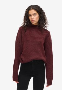 Burgundy kötött pulóver bordás textúrával, magas nyakú és laza fazonú. Fekete, magas derekú farmerral párosítva, széles ujjaival.