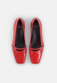 Zapatos de charol rojo con punta cuadrada, tacón plano y una correa con una hebilla de plata. El interior es negro con detalles del logo.