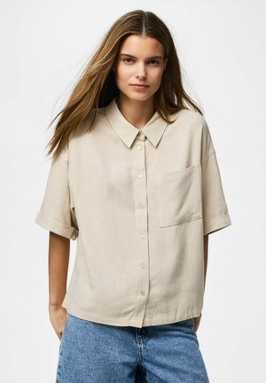 Jeune femme aux longs cheveux portant une chemise beige à manches courtes boutonnée et un jean bleu sur un fond blanc uni.