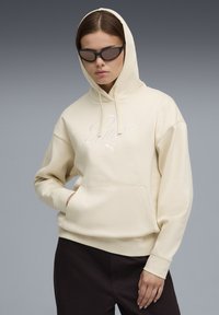 Krémová mikina s kapucňou so prednou kapsou a vyšitým logom "Puma". Model má na sebe čierne slnečné okuliare na pozadí s gradientom.