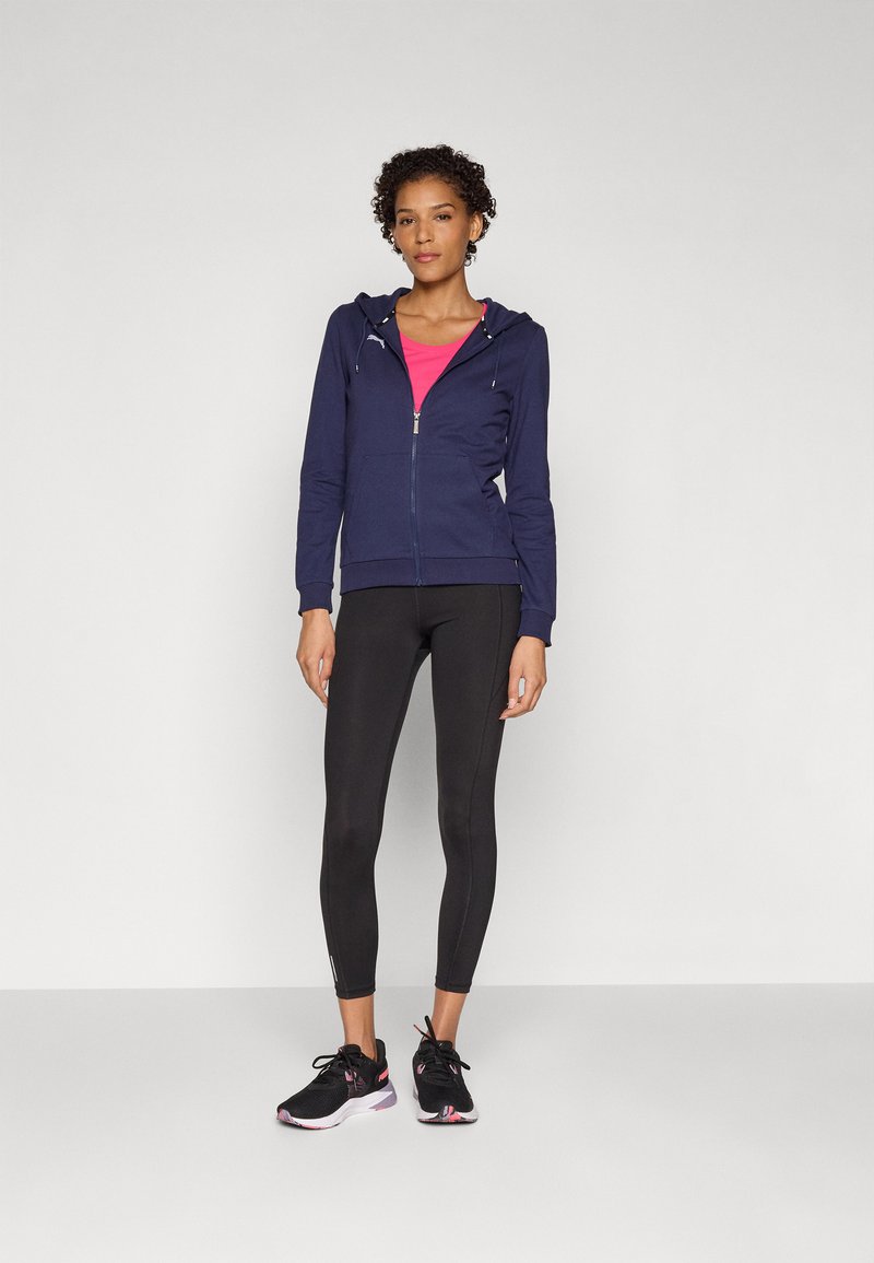 Felpa con zip della marina, con coulisse, top rosa sotto, leggings neri e scarpe da corsa nere con dettagli e tessuto testurizzato.