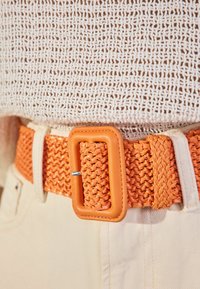 Båret orange bælte med et rektangulært læderspænde, der har en tekstureret overflade og glatte detaljer, kombineret med lyse bukser.