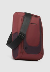 Bolso de hombro rojo y púrpura oscuro hecho de tela texturizada, con una correa de hombro acolchada y compartimentos con cremallera. Forma rectangular.