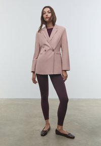 Donna in piedi su un pavimento di cemento, che indossa una giacca beige a doppio petto, leggings prugna e ballerine marroni scure su uno sfondo bianco.