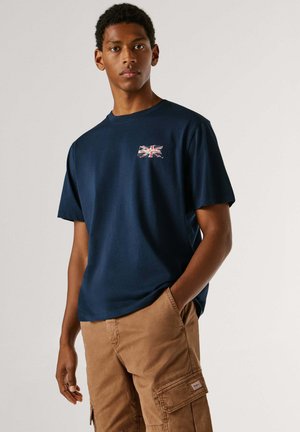 Junger Mann trägt ein marineblaues T-Shirt mit kleinem Union-Jack-Flaggendruck und hellbraune Cargo-Shorts, steht mit einer Hand in der Tasche.
