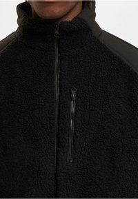 Schwarze Fleecejacke mit durchgehendem Reißverschluss, hohem Kragen und einer Reißverschlusstasche auf der Brust, getragen von einer Person mit dunkler Hautfarbe.