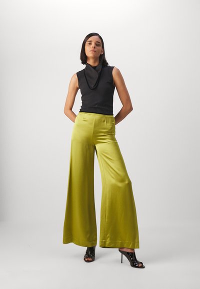Zwart mouwloos topje met een capuchonnek, gecombineerd met hoge taille, wijde groene satijnen broeken. Zwarte open-taille hoge hakken accentueren de outfit.
