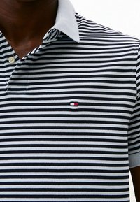 T-shirt polo avec des rayures horizontales bleu marine et blanches, un col gris clair et un petit logo brodé en rouge et bleu sur la poitrine.