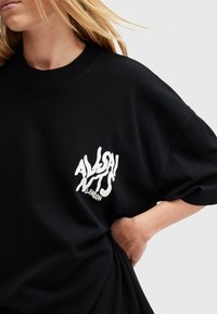 Černé oversize tričko vyrobené z bavlny. Na levé části hrudníku má bílý grafický potisk "ALLSAINTS LONDON". Krátké rukávy.