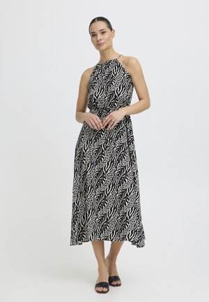 Moteris stovi vilkėdama juodai baltą be rankovių midi su gyvūnų raštu ir juodus atvirus aukštakulnius, fone yra paprasta balta spalva.