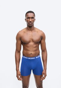 Μυώδης άνδρας στέκεται με το πρόσωπο προς τα εμπρός φορώντας μπλε μποξεράκια Björn Borg σε απλό γκρι φόντο.