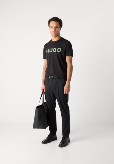 Черен т-shirt с bold зелено "HUGO" принт, съчетан с черни панталони и черни маратонки. Държи черна чанта. Изцяло прост дизайн.