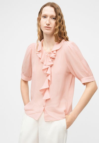 Femme aux cheveux ondulés portant un chemisier rose clair à volants avec des manches courtes bouffantes et un pantalon blanc, mains dans les poches, fond neutre.