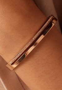 Roze gouden armband heeft een slank ontwerp met een dunne, rechte band en een rij kleine heldere stenen die langs één rand zijn ingelegd.