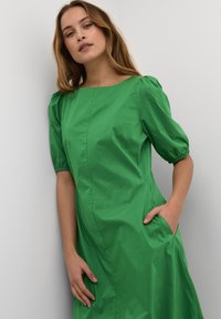 Rochie verde cu mâneci bufante, confecționată dintr-un material ușor, având decolteu pătrat și buzunare laterale, cu o textură netedă și mată.