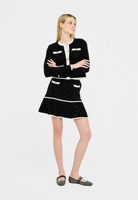 Zwart-wit tweedelig ensemble met een nauwsluitende cardigan en een uitlopende rok, voorzien van zakken met knopen en een gespikkelde textuur.