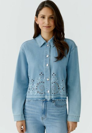 Jonge vrouw met een lichtblauw spijkerjack met bloemige uitsnijdingen en bijpassende spijkerbroek, staand tegen een effen achtergrond.