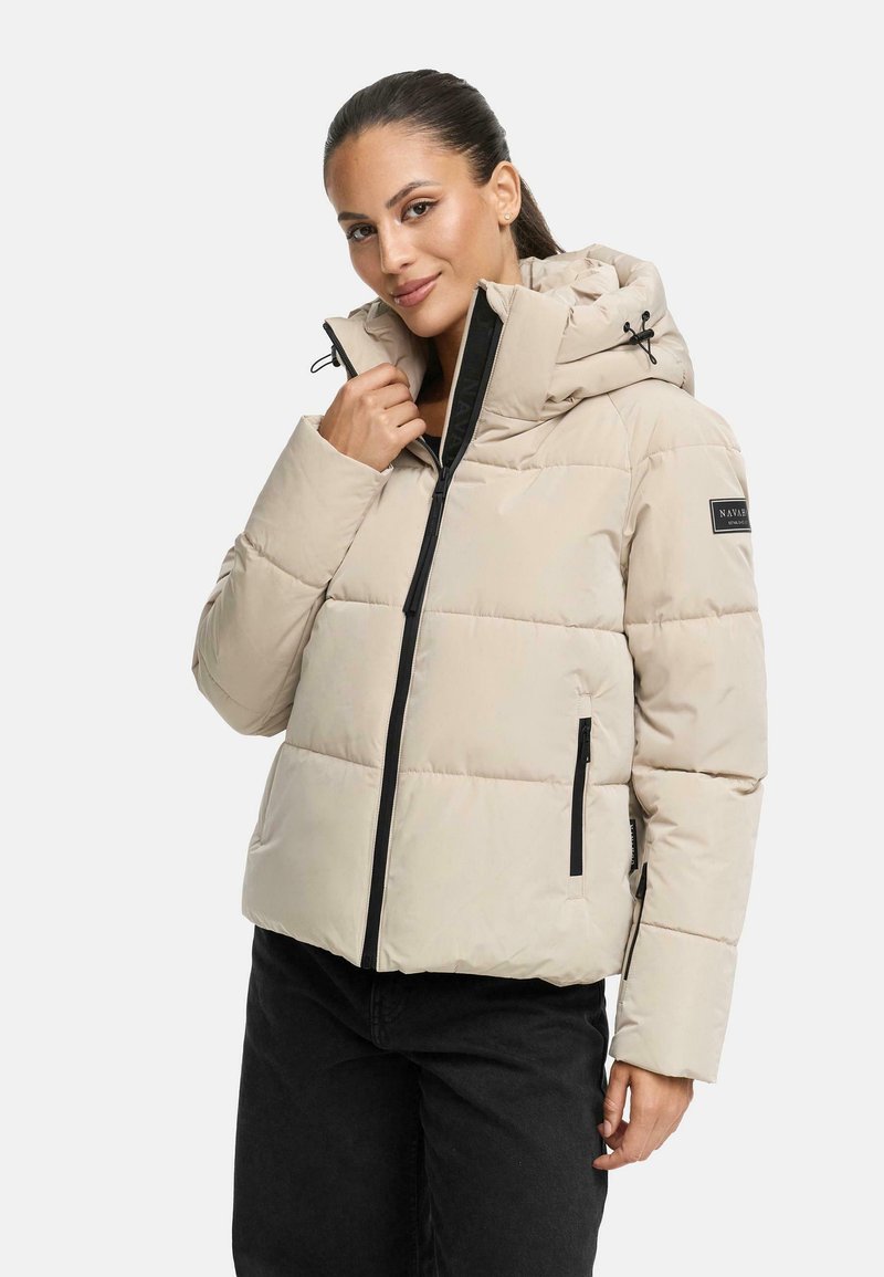 Veste matelassée beige avec capuche, dotée de fermetures éclair et de détails noirs. Elle présente un design en matelassage et une patch logo sur la manche.