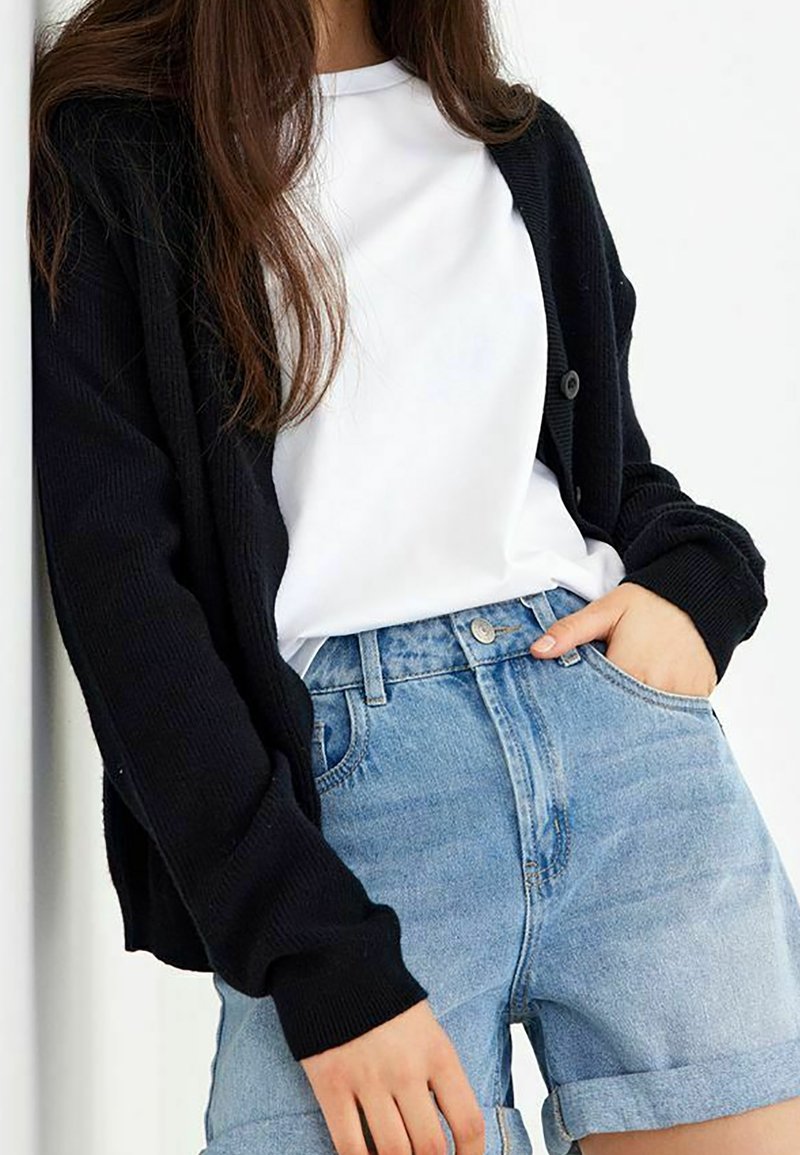 Cardigan noir en tricot avec texture côtelée, porté sur un T-shirt blanc et un short en denim bleu clair avec un ourlet replié. Boutons sur le cardigan.
