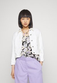 Casaco de ganga branco com botões de metal, blusa estampada de flores em preto, calças roxas de cintura alta com uma textura suave. Cabelo curto e escuro.