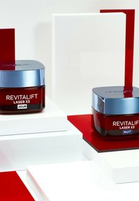 Des bocaux rouges étiquetés "Revitalift Laser X3 Jour" et "Nuit" avec des couvercles bleu métallique exposés sur des blocs géométriques blancs et rouges.