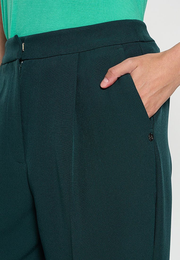 Donkergroene broek van gladde stof, met een zijzak en een kleine metalen accent. Het ontwerp is op maat gemaakt met een lichte drape.