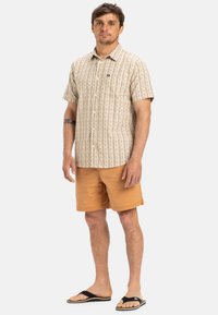 Camisa de botones de manga corta beige con rayas verticales marrón claro, combinada con pantalones cortos marrón claro y chanclas negras.
