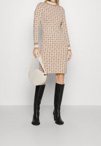 Robe longue à manches longues à motif marron avec un col montant, associée à des bottes noires montantes jusqu'aux genoux et un sac à épaule beige clair.