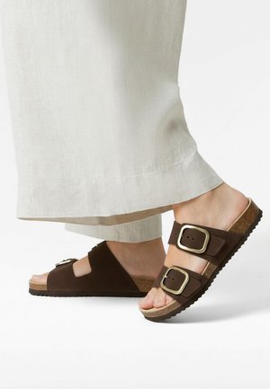 Smeđe antilop natikače na navlačenje s dvostrukim podesivim kopčama, pluto uloškom i teksturiranim gumenim potplatom, u kombinaciji sa svijetlim lanenim hlačama.