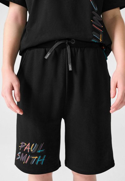 Shorts noirs avec le texte multicolore "Paul Smith" sur la jambe gauche, taille élastique avec cordon de serrage, portés par une personne les mains détendues le long du corps.