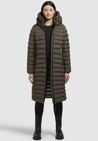 Olive green long puffer coat with a hood, featuring a quilted pattern, side zip pockets, and a slim fit, worn over black clothing.