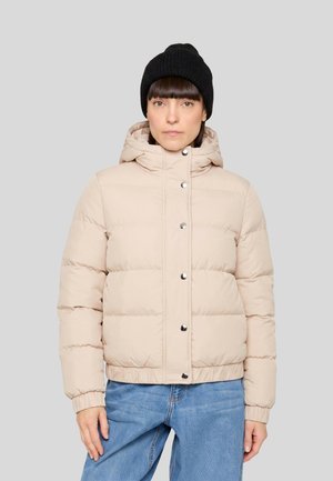 Femme portant une veste matelassée beige avec des boutons-pression, un jean bleu et un bonnet en tricot noir, se tenant devant un fond gris uni.