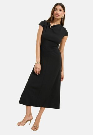 Donna che indossa un vestito midi nero con maniche a cappuccio e una chiusura dorata sullo scollo, orecchini a cerchio dorati, braccialetto e sandali dorati con tacco.